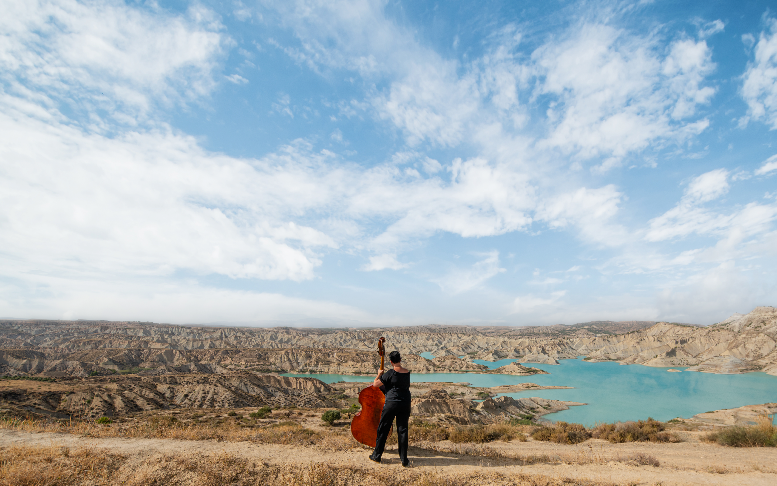 Músico con contrabajo ante un embalse rodeado de paisaje árido